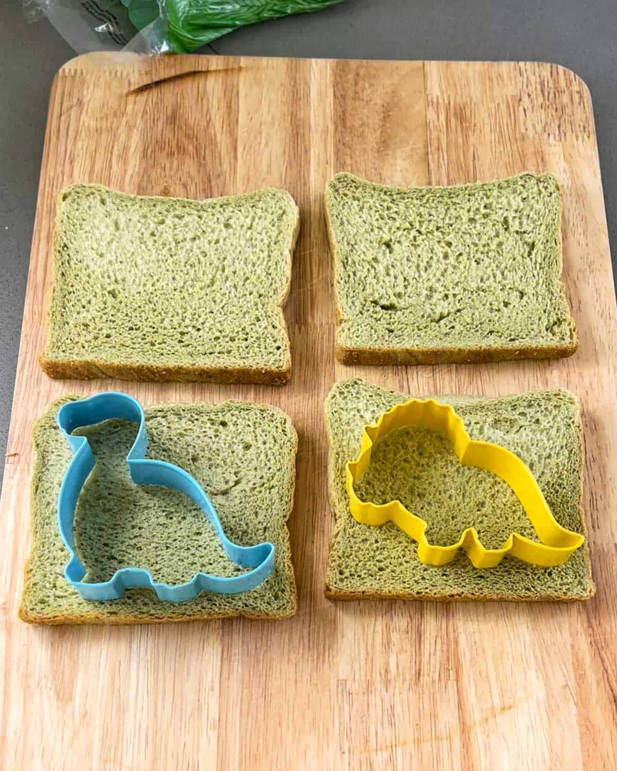 Dinosaur cookie cutters on pieces of spinach bread on a wooden chopping board.