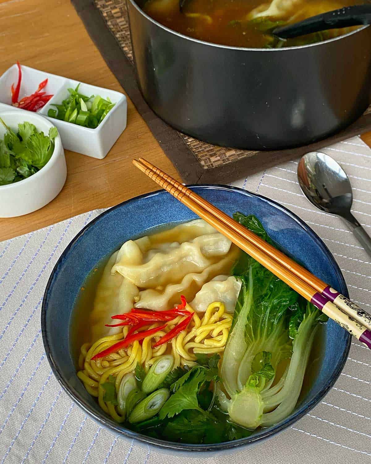 A bowl of Dumpling Noodle Soup surrounded by bowls of garnish and chopsticks.