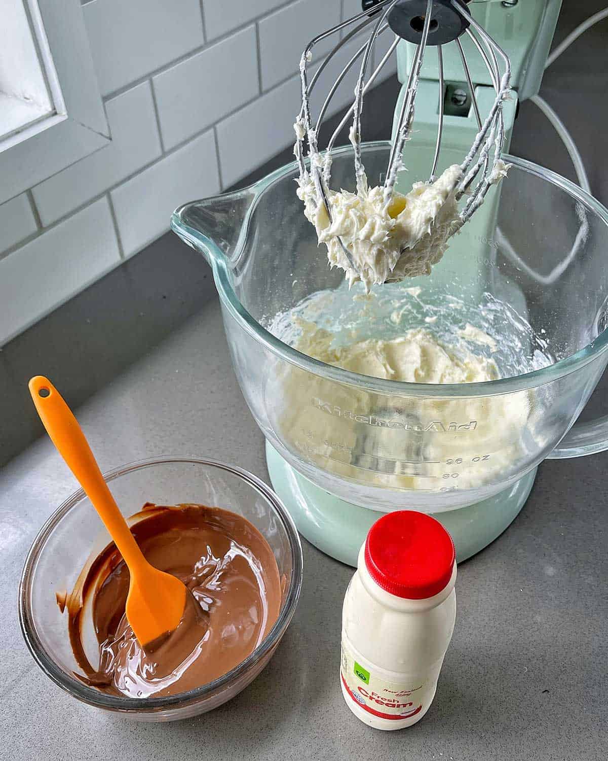 A mixer with whipped cream in it, a bowl of melted chocolate and a bottle of cream sitting on a bench.