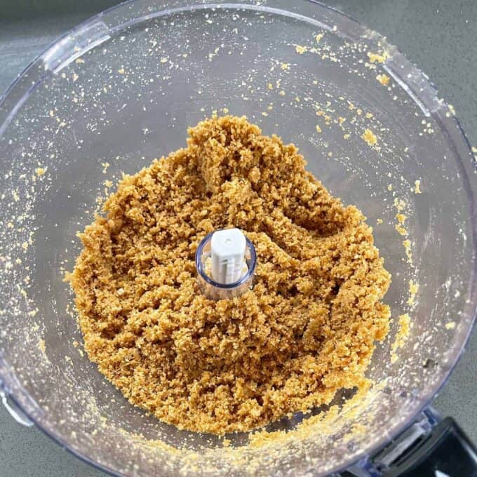 A food processor contains a crumbly mixture, likely a blend of crushed biscuits or cookies, forming a grainy dough-like consistency perfect for making Easter Cheesecake Cups.
