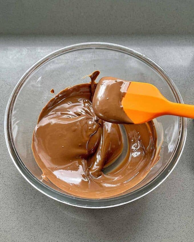 A glass bowl with smooth, melted chocolate being stirred by an orange spatula on a gray countertop, perfect for creating delightful Easter Cheesecake Cups.