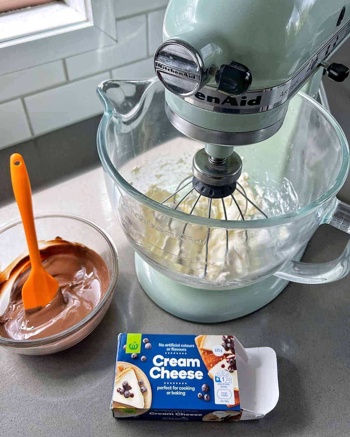 A stand mixer with cream cheese in it next to a bowl of melted chocolate and a packet of cream cheese.