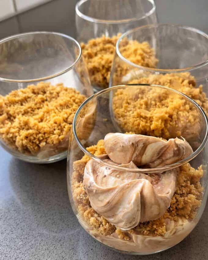Four glasses filled with crumbled biscuit base and chocolate mousse, elegantly capturing the essence of Easter Cheesecake Cups, rest on a gray countertop.