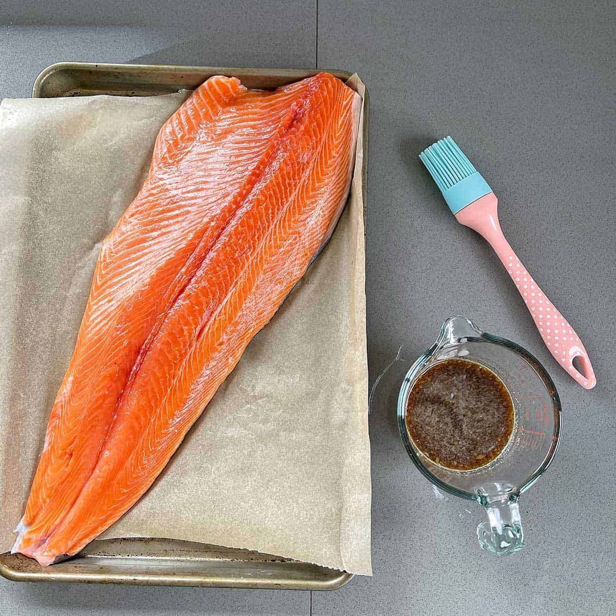 A whole fillet of salmon on a lined baking tray next to a jar of glaze and a brush.