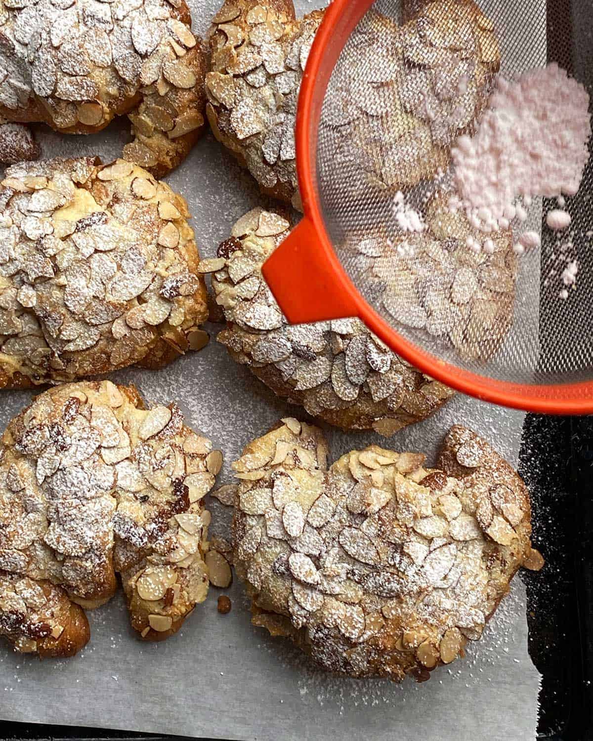 Almond Croissants being sprinkled with icing sugar from a red sieve. 