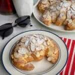 An Almond Croissant on a white plate next to some sunglasses, a mug and a plate of almond croissants.