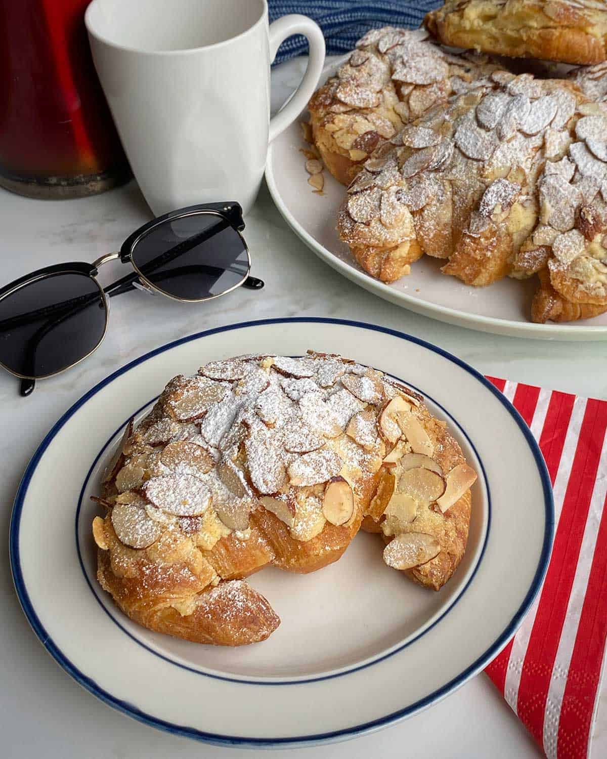 An Almond Croissant on a white plate next to some sunglasses, a mug and a plate of almond croissants.