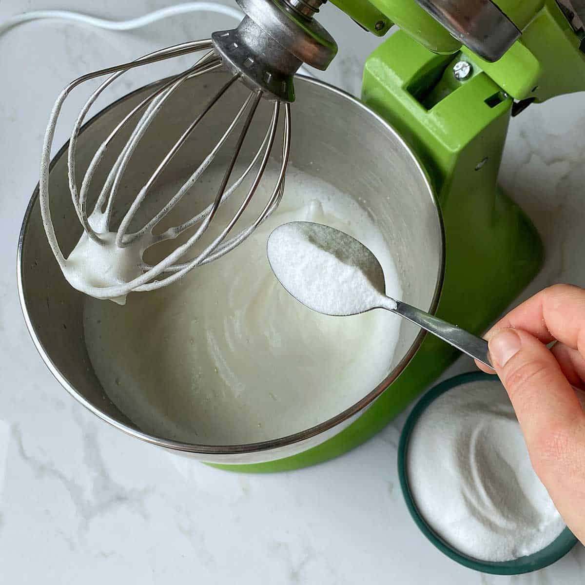Sugar being added to whipped egg white in the bowl of a stand mixer.