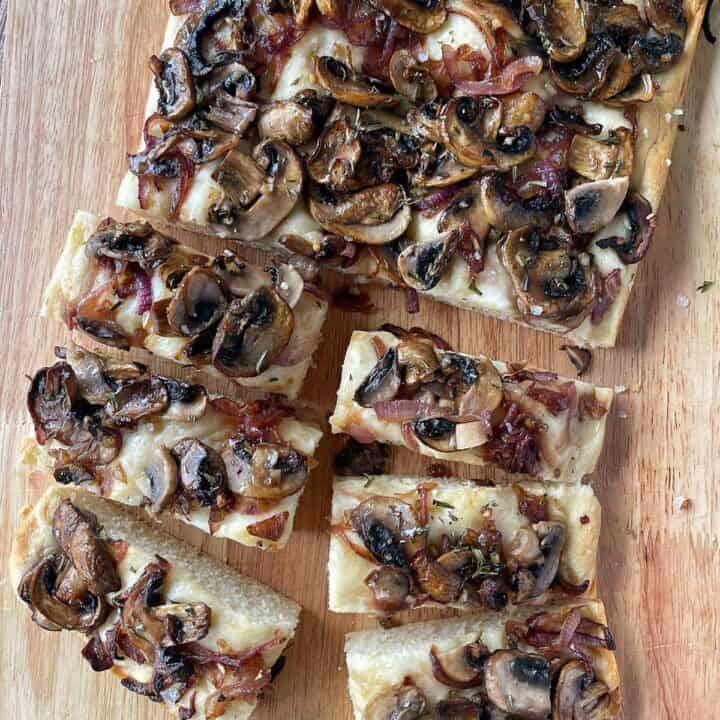 A sliced loaf of mushroom and red onion focaccia sitting on a wooden chopping board.