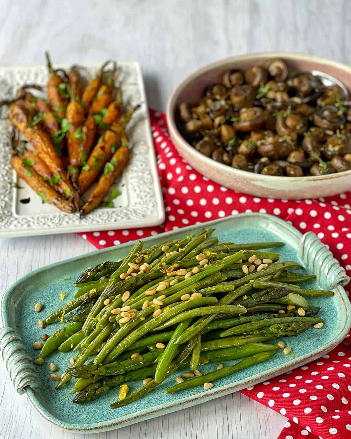 Roasted carrots, sauteed greens and baked mushrooms sitting on individual platters on a red tablecloth on a white table.