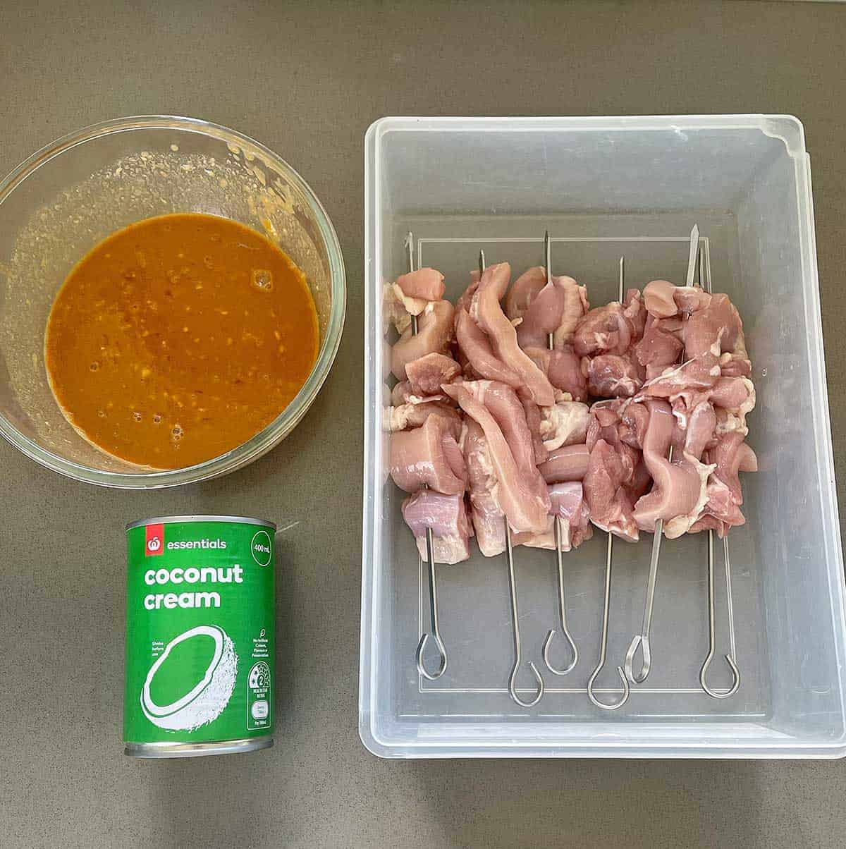 Satay marinade in a glass bowl, a can of coconut milk and a container of raw chicken skewers sitting on a grey bench.
