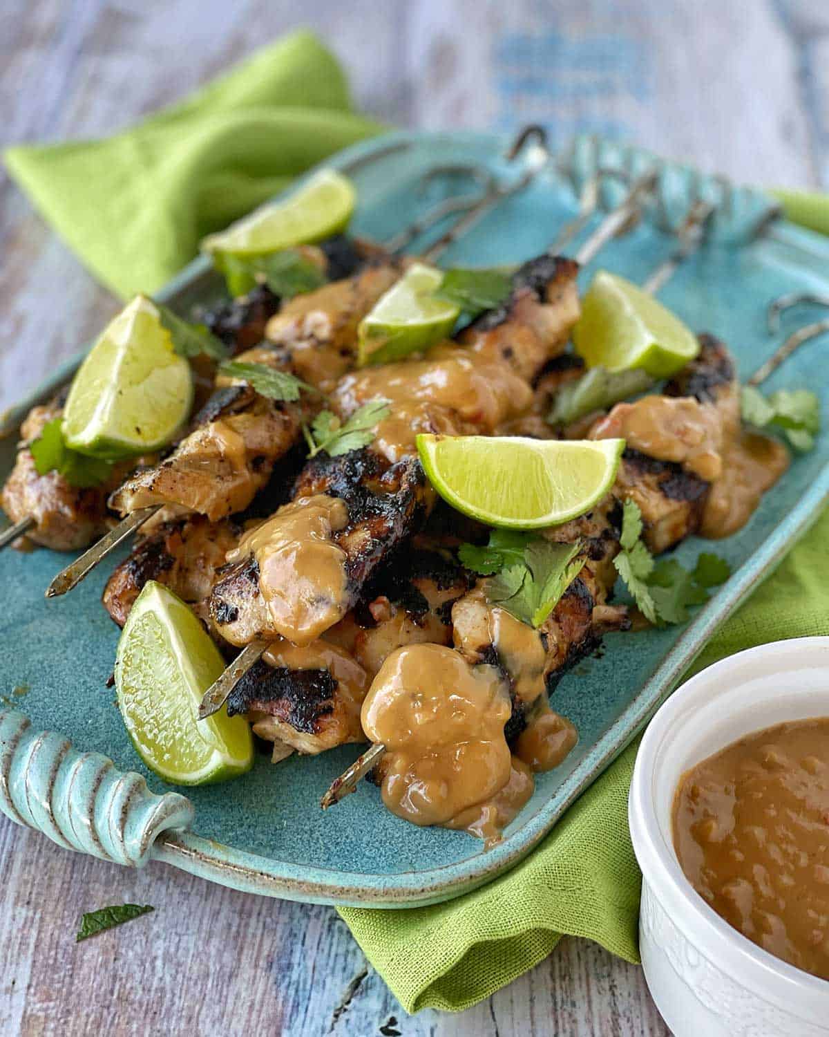 BBQ Satay Chicken Skewers  on a blue platter piled up with lime wedges and coriander.