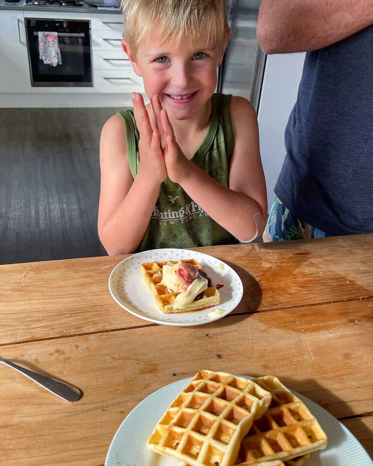 A smiling young boy sitting at a wooden table with a plate of waffles in front of him.