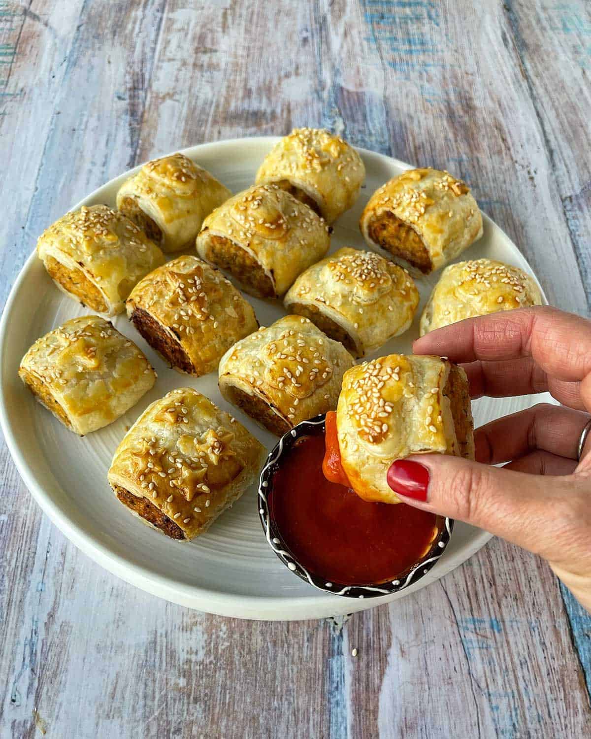 A hand holding a sausage roll, dipped in tomato sauce taken from a platter of sausage rolls.