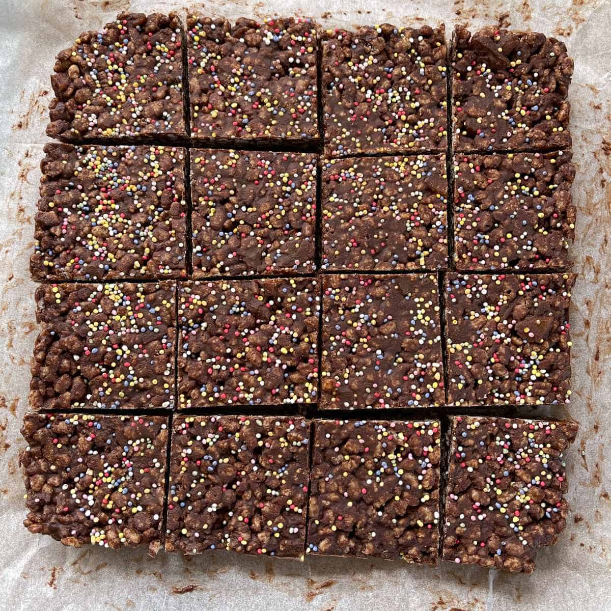 Chocolate rice bubble slice with sprinkles cut into 16 pieces on top of baking paper