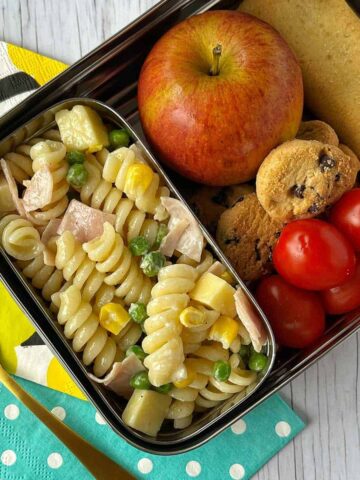 Kids' Pasta Salad in a container in a stainless steel lunchbox filled with fruit and biscuits.