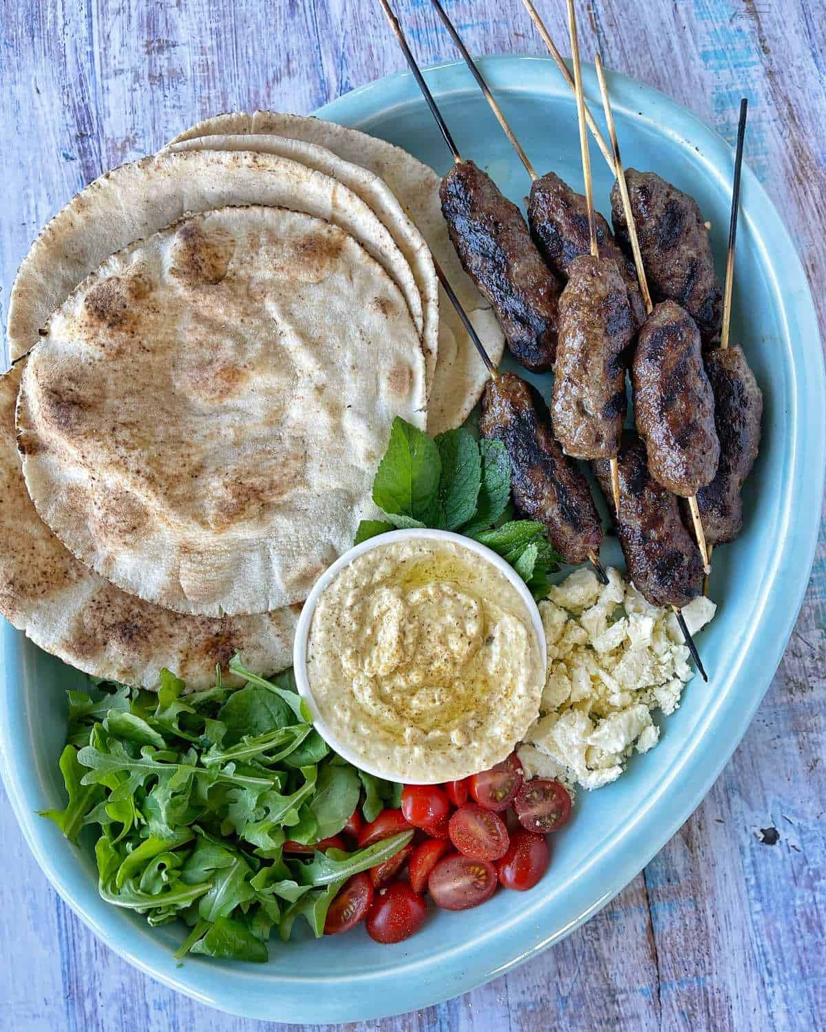 Lamb koftas and vegetables and wraps on a blue platter.