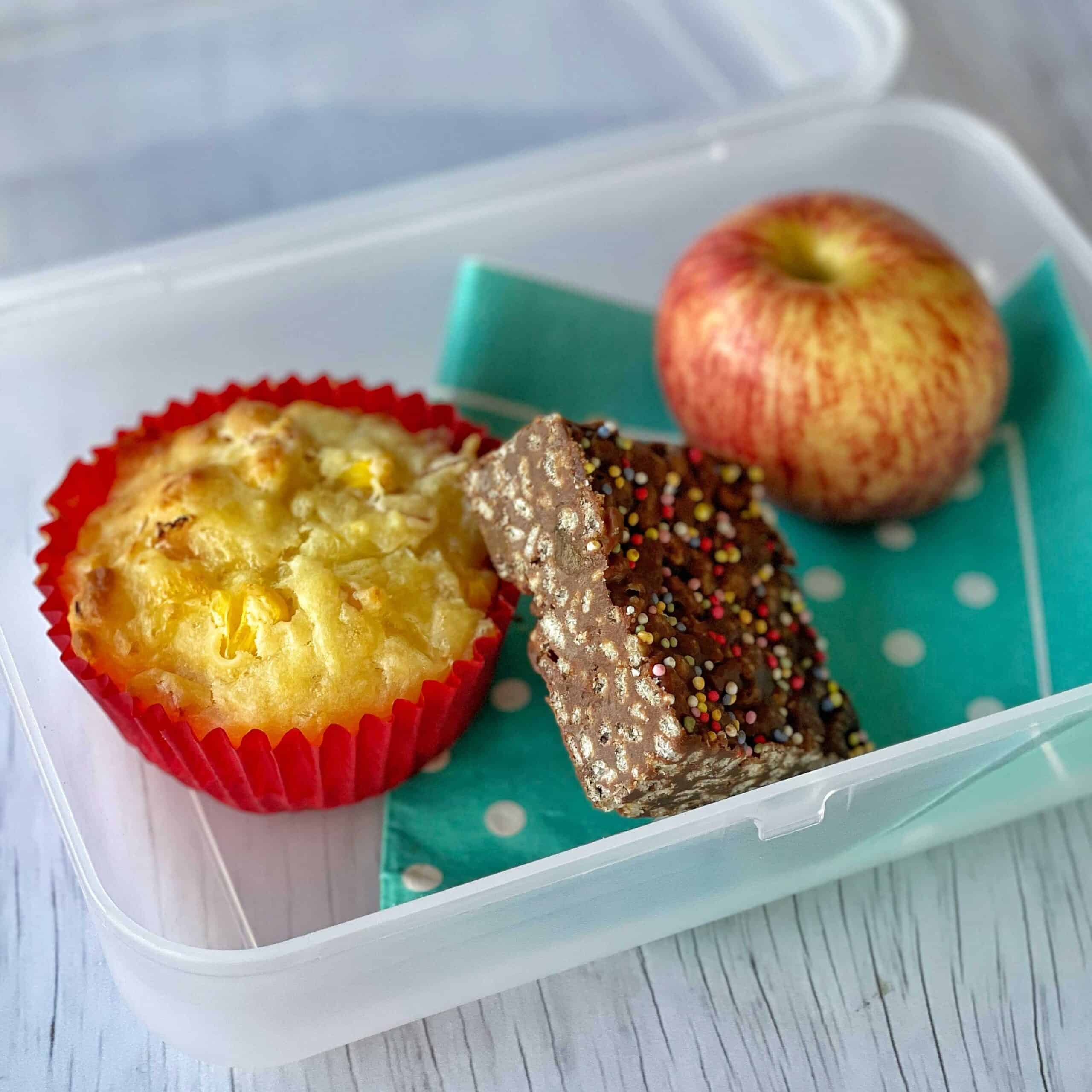 A savoury muffin, a piece of chocolate slice and an apple sitting in a plastic lunchbox.