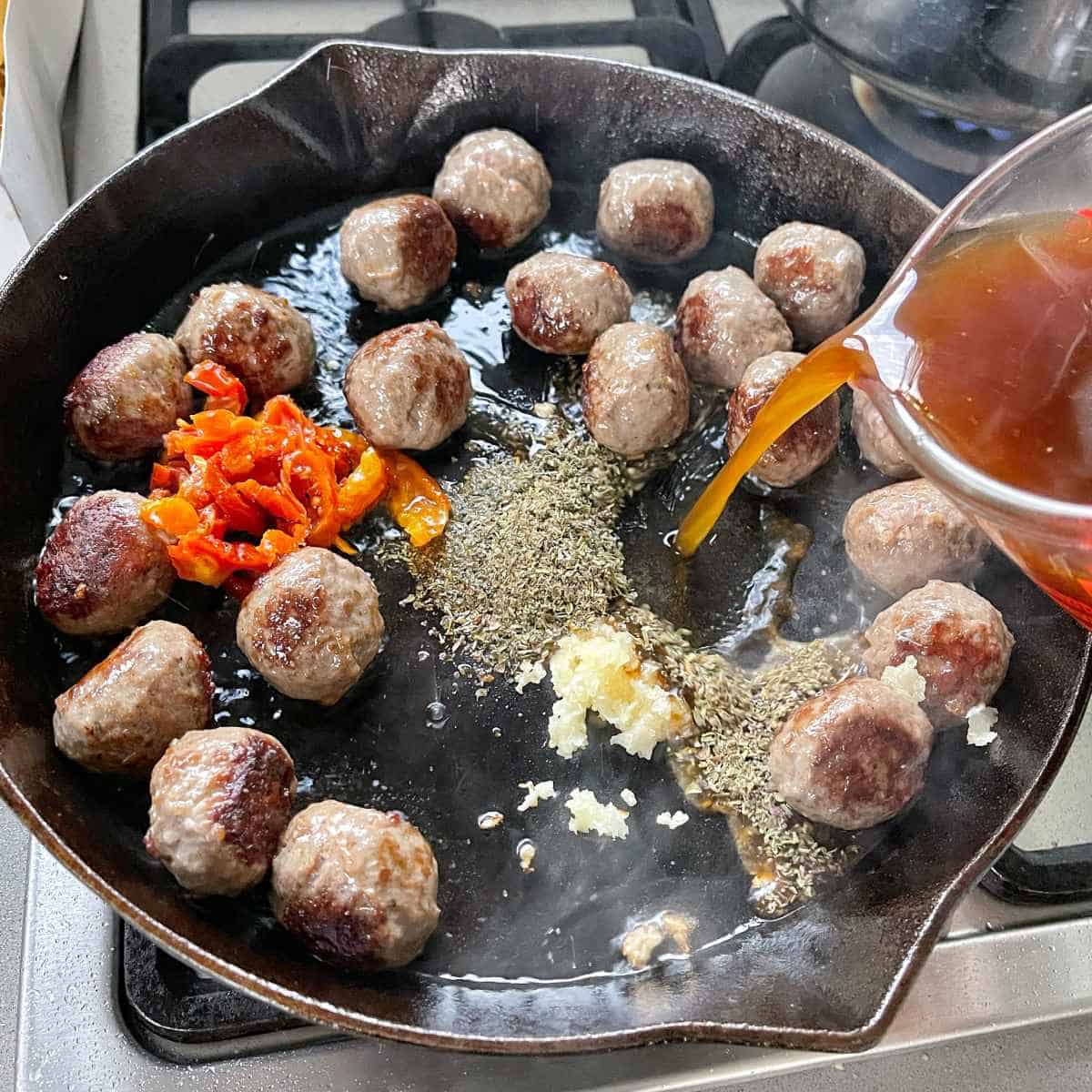 Slightly cooked meatballs frying in a medium frypan, herbs, garlic, sundried tomato's have been added. Beef stocked is being poured into pan from a glass jug.