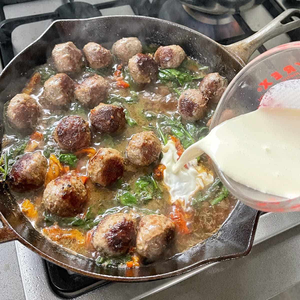 Tuscan meatballs simmering over a medium heat with cream being added from a small glass jug