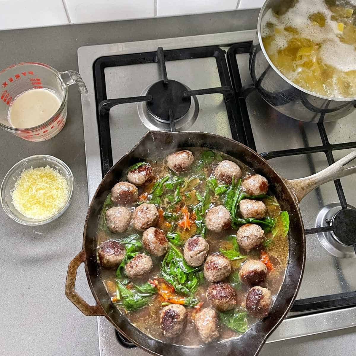 Birds eye view of Tuscan meatballs simmering over a low heat on the element. Saucepan of boiling water and pasta cooking with a glass jug of cream and small glass bowl of cheese sitting on a grey bench top