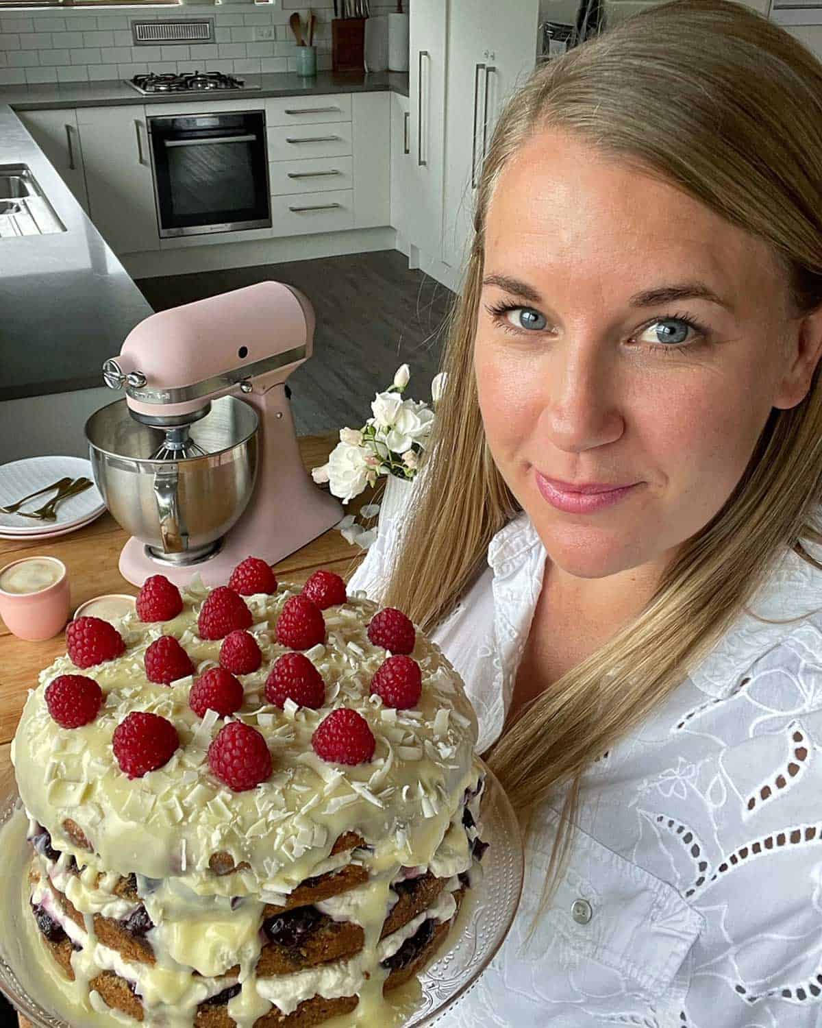 A woman holding a White Chocolate Cherry Cake with her kitchen behind her.