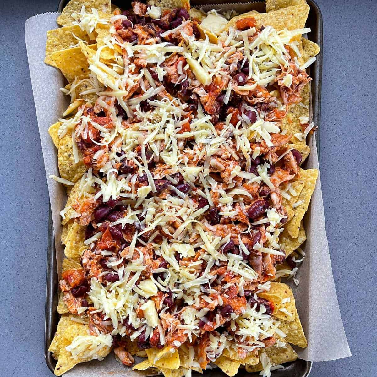 Cheesy chicken nachos assembled on a baking tray with grated cheese on top, ready to go into the oven on grill