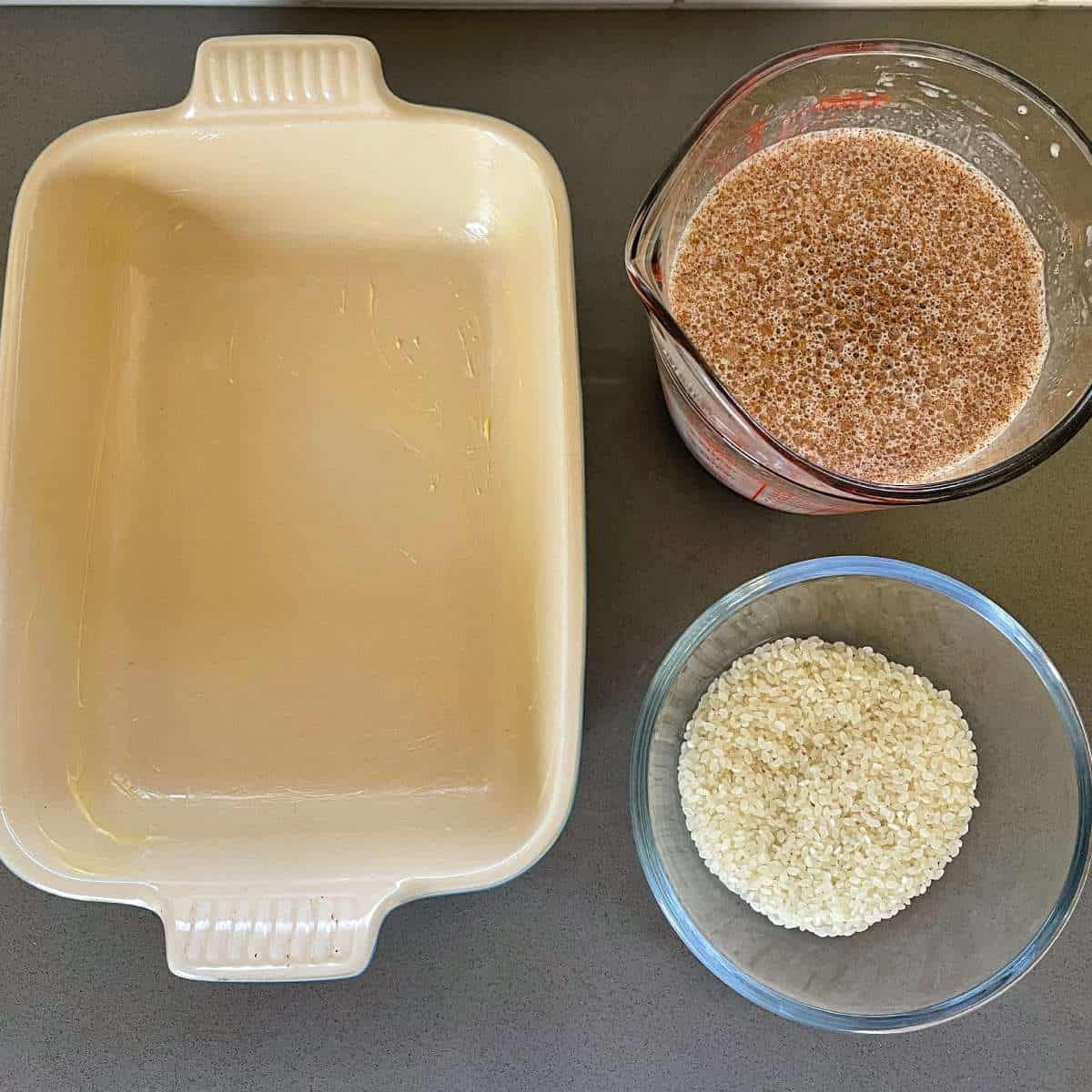 Two different dishes filled with the wet and dry ingredients for a rice pudding beside a ovenproof dish sitting on a grey bench