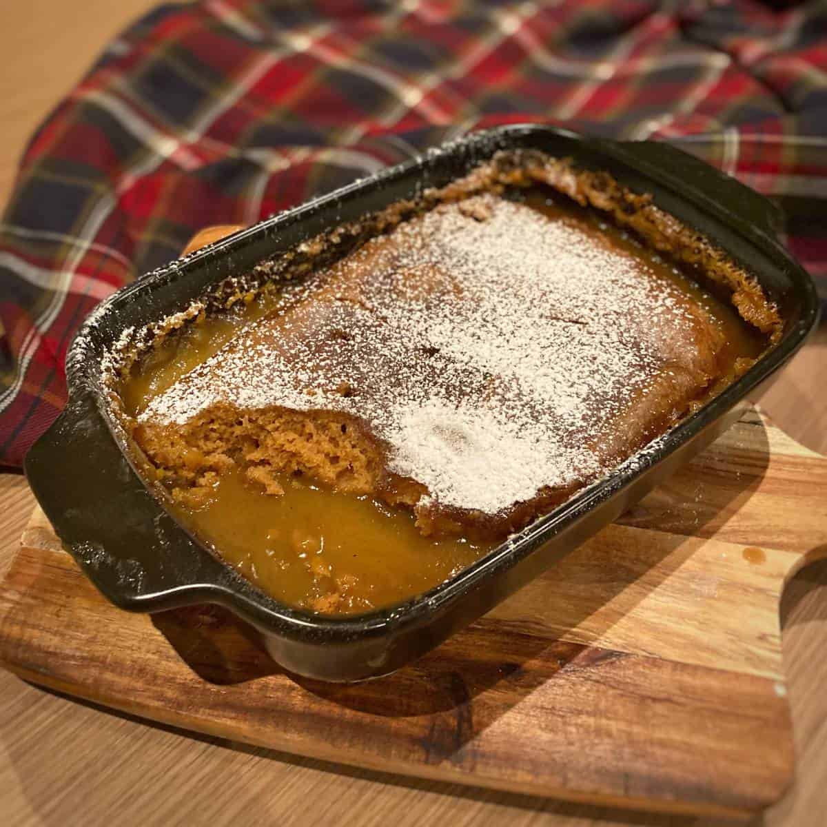 Gingerbread pudding sitting in the dish it was cooked in on top of a wooden chopping board