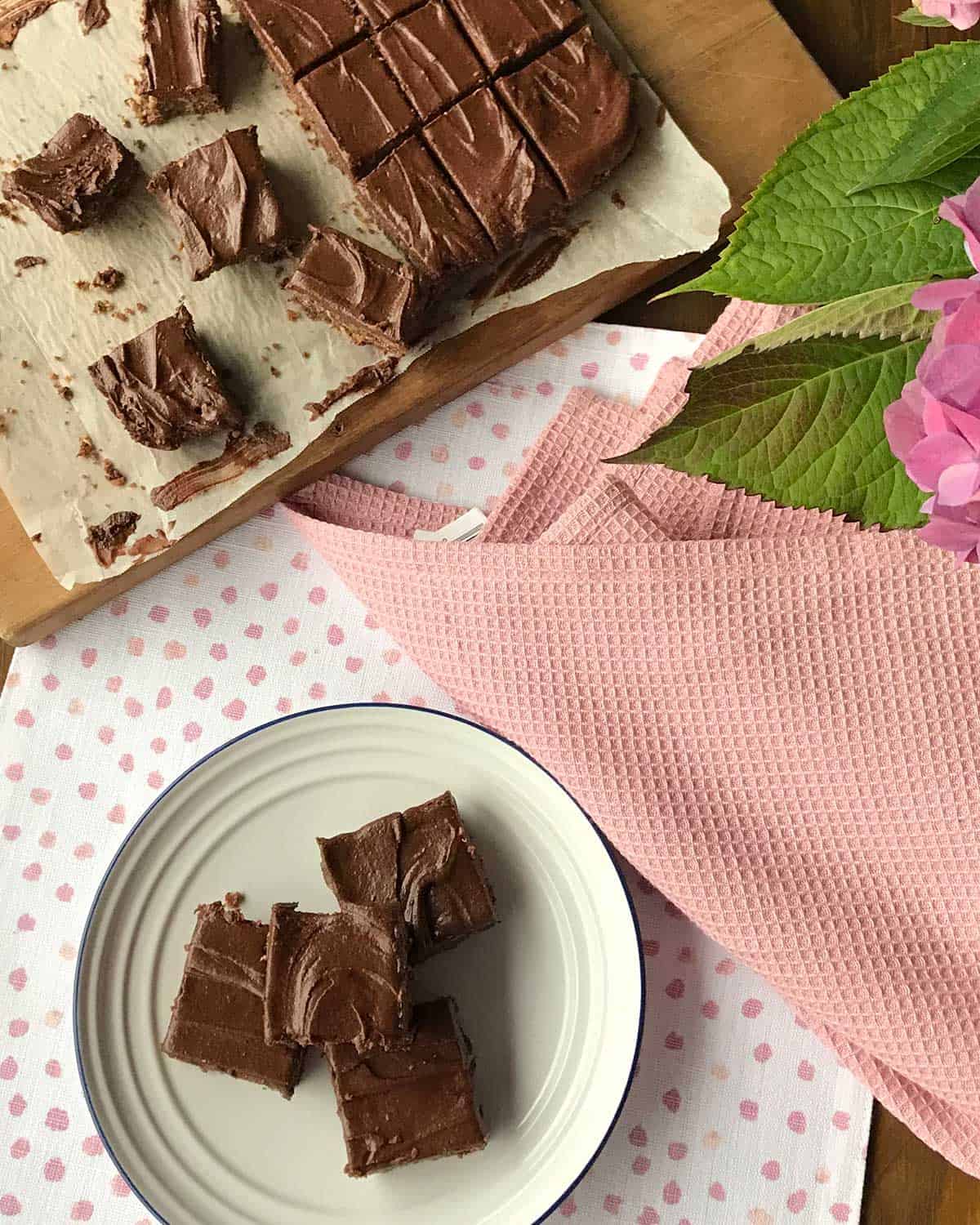 Three pieces of chocolate weetbix slice on a white plate next to a chopping board with the rest of the slice.