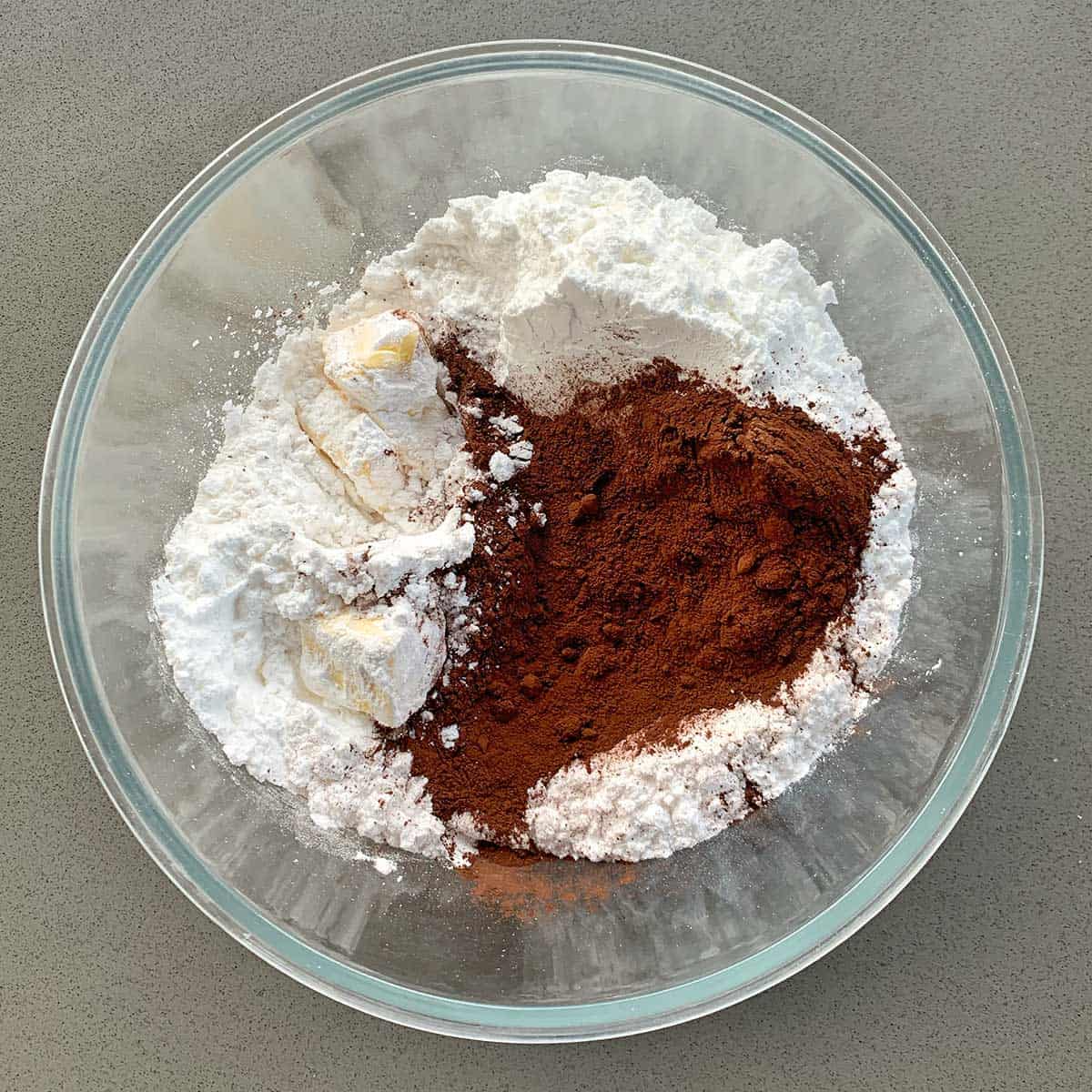 Icing sugar, butter and cocoa in the base of a glass bowl on a grey bench.