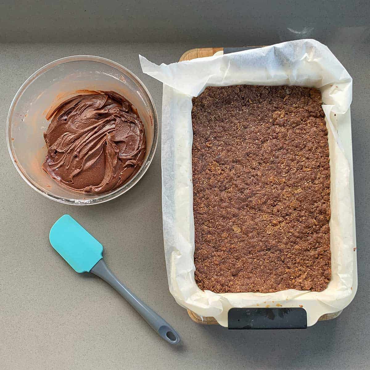 Chocolate icing, a spatula and the cooked base of Chocolate Weetbix Slice sitting next to each other on a grey bench.