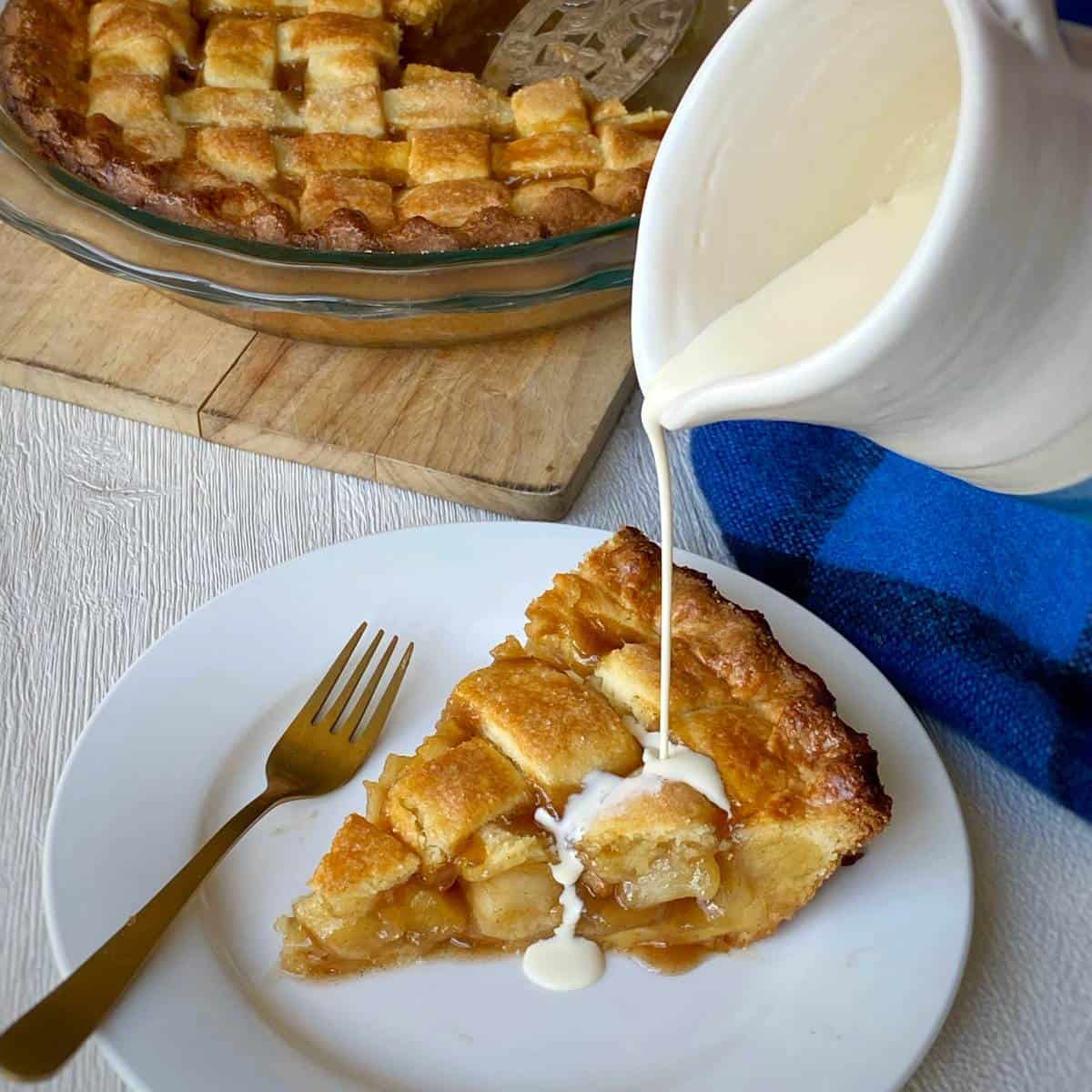 A slice of apple pie on a white plate with cream being poured over it