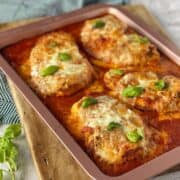 Baked chicken parmigiana sitting in a dusty pink oven proof on top of a wooden chopping board.