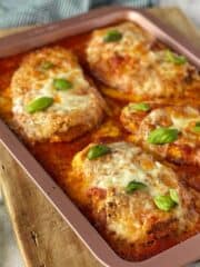 Baked chicken parmigiana sitting in a dusty pink oven proof on top of a wooden chopping board.