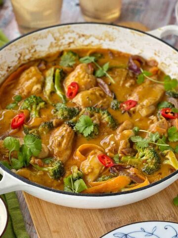 Cooked chicken satay yellow curry in a large white round oven dish sitting on a wooden chopping board