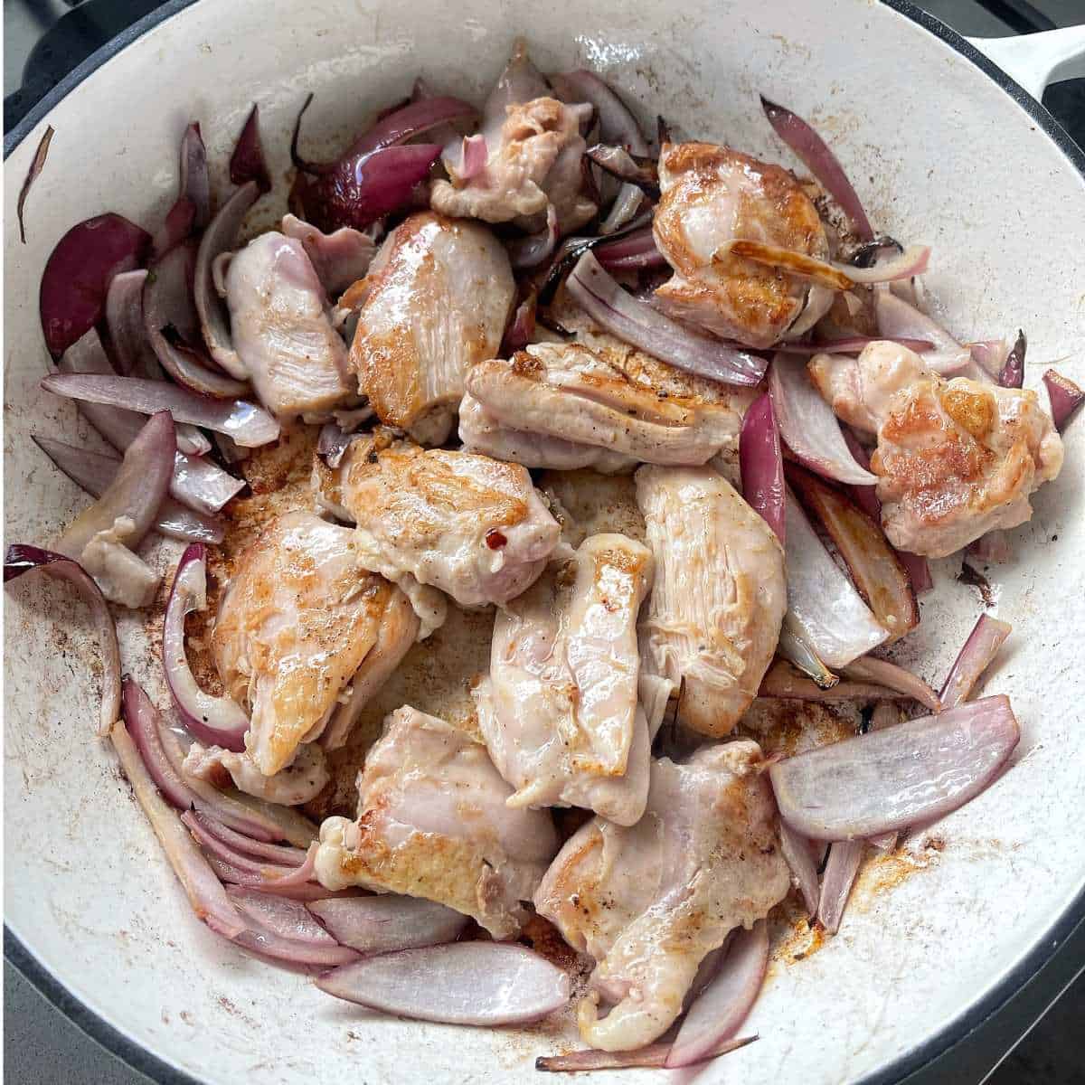 Red onion and sliced chicken pieces of chicken frying over a hob in large white, round oven dish