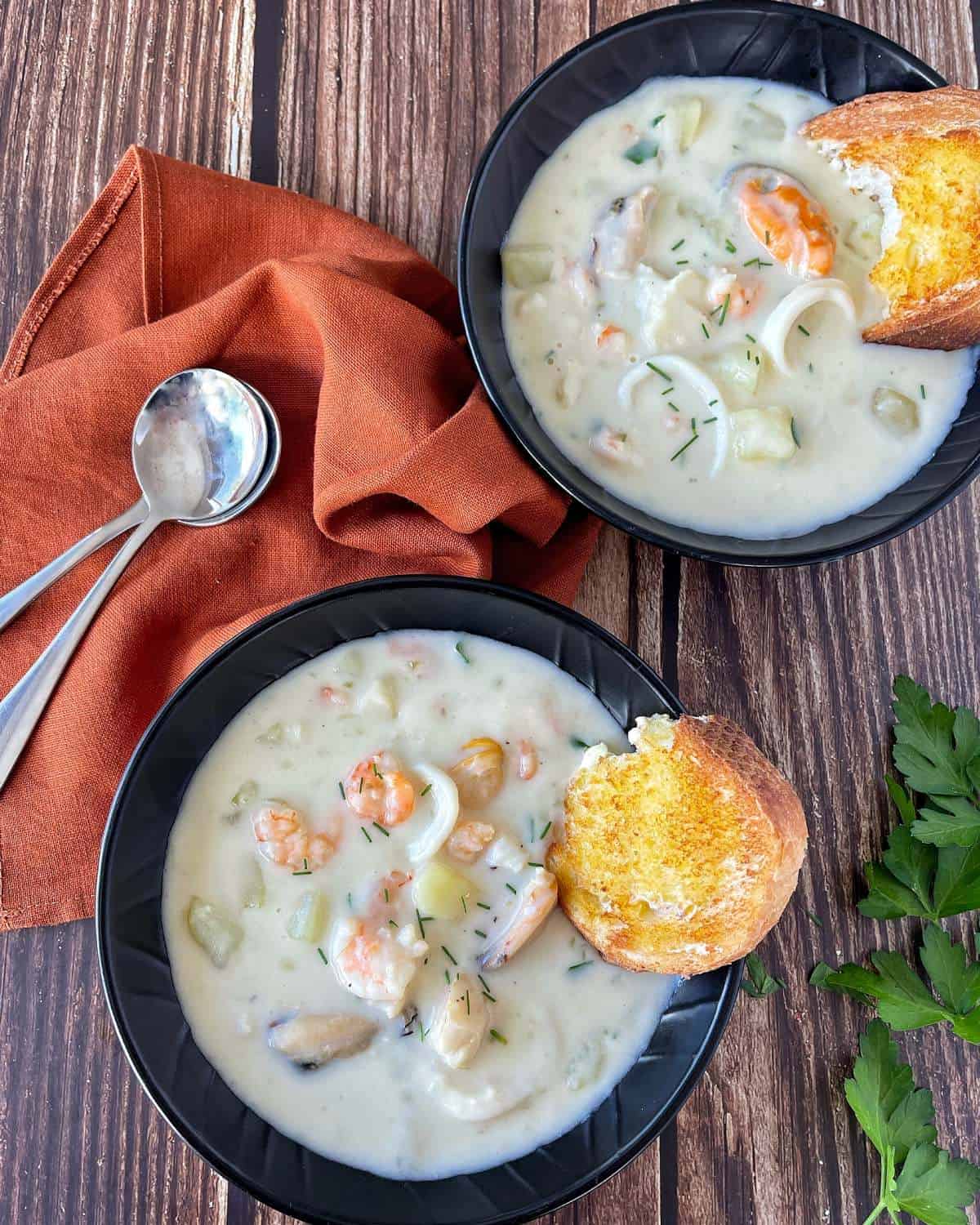 Two black bowls of seafood chowder served with a slice of toasted tiger loaf, with butter on a wooden bench top