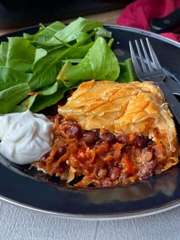 A slice of veg mix pie served with a green salad and a blob of sourcream on a navy blue plate.