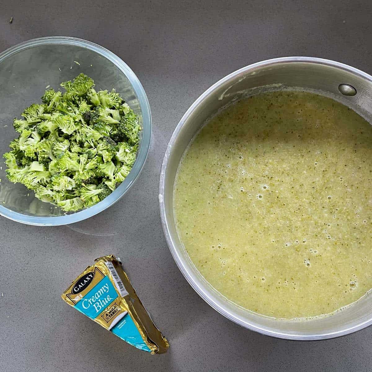 Cooked blue cheese and broccoli soup in a saucepan with a small glass bowl of broccoli pieces and a packet of blue cheese sitting on a grey bench top