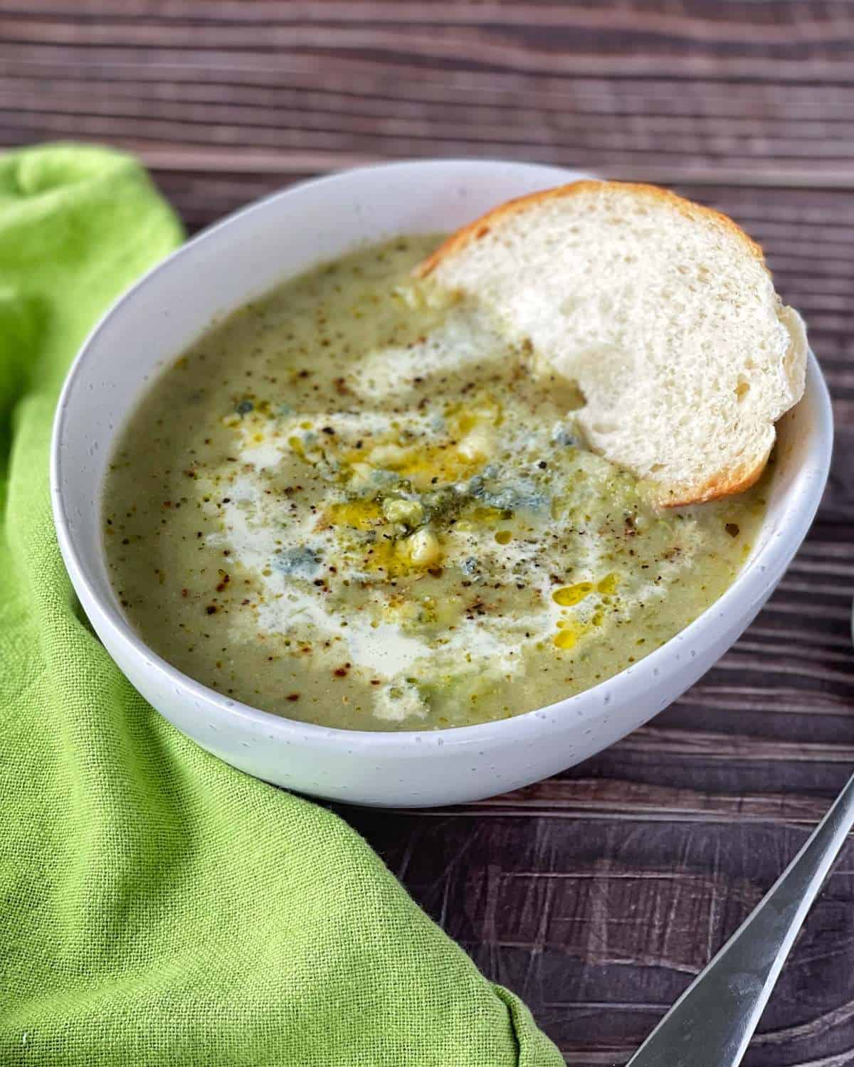 Blue cheese and broccoli soup served in a white soup bowl with a slice of fresh bread, a green napkin to the side on a wooden table