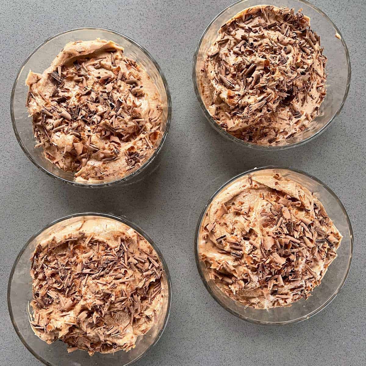 Chocolate mousse with grated chocolate on top in glass bowls on a grey bench.