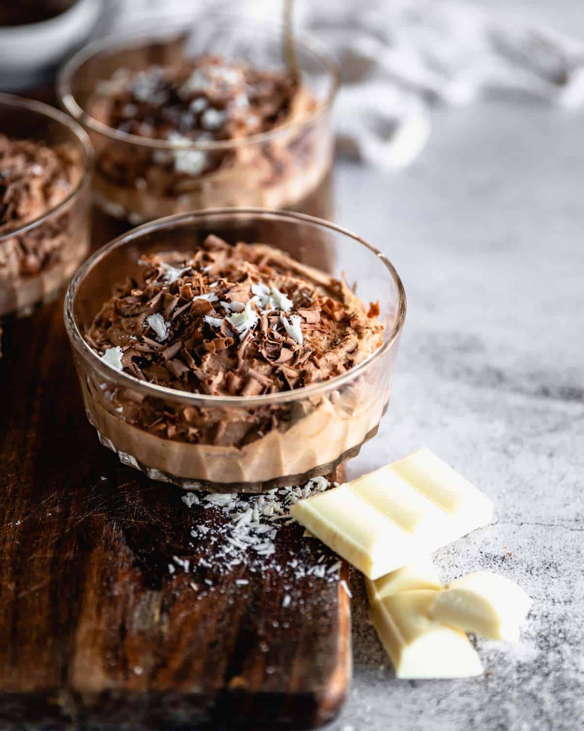 A close up of one serving of chocolate mousse with shredded coconut and chocolate on top with some pieces of white chocolate to the side on a wooden chopping board