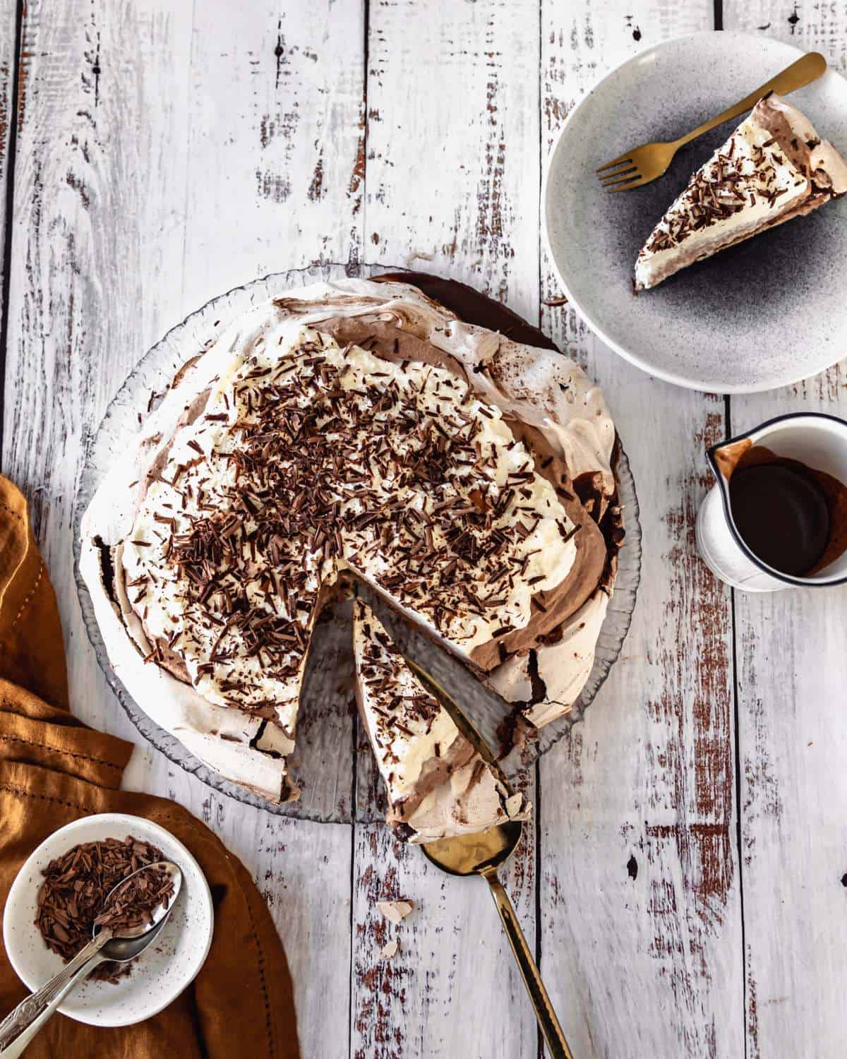 Cooked and assembled chocolate pavlova with a slice removed and served in a grey dessert bowl. Small jug of chocolate sauce and grated chocolate to the side