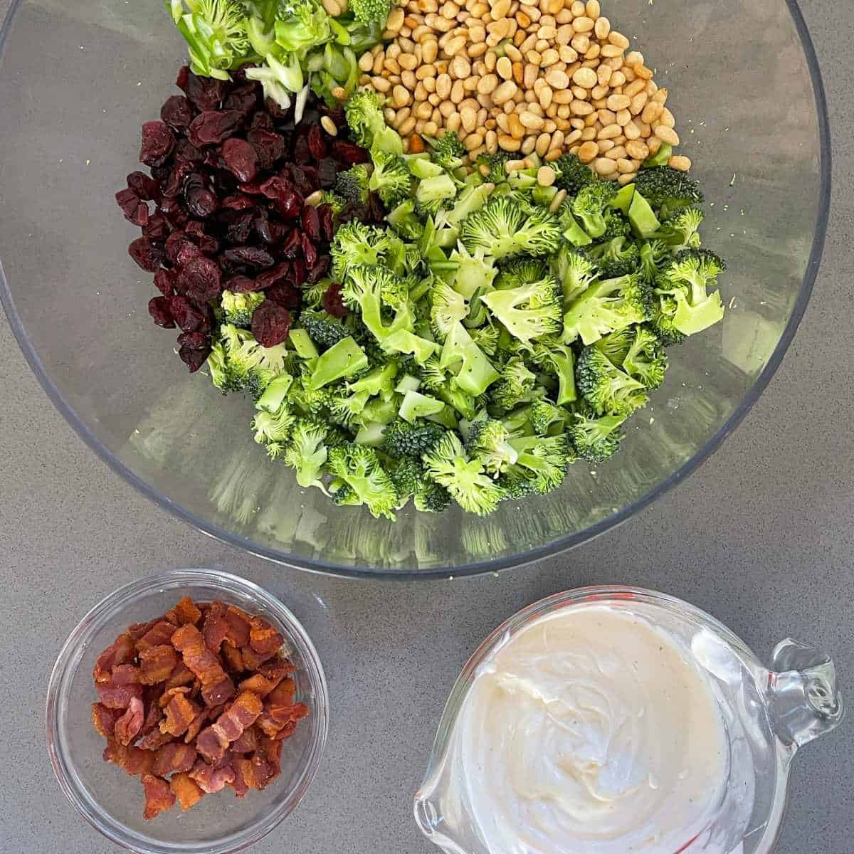 Three bowls on a grey bench top. One with toasted pine nuts, cranberries and cut raw broccoli, the second with fried bacon pieces and finally the dressing for raw broccoli salad