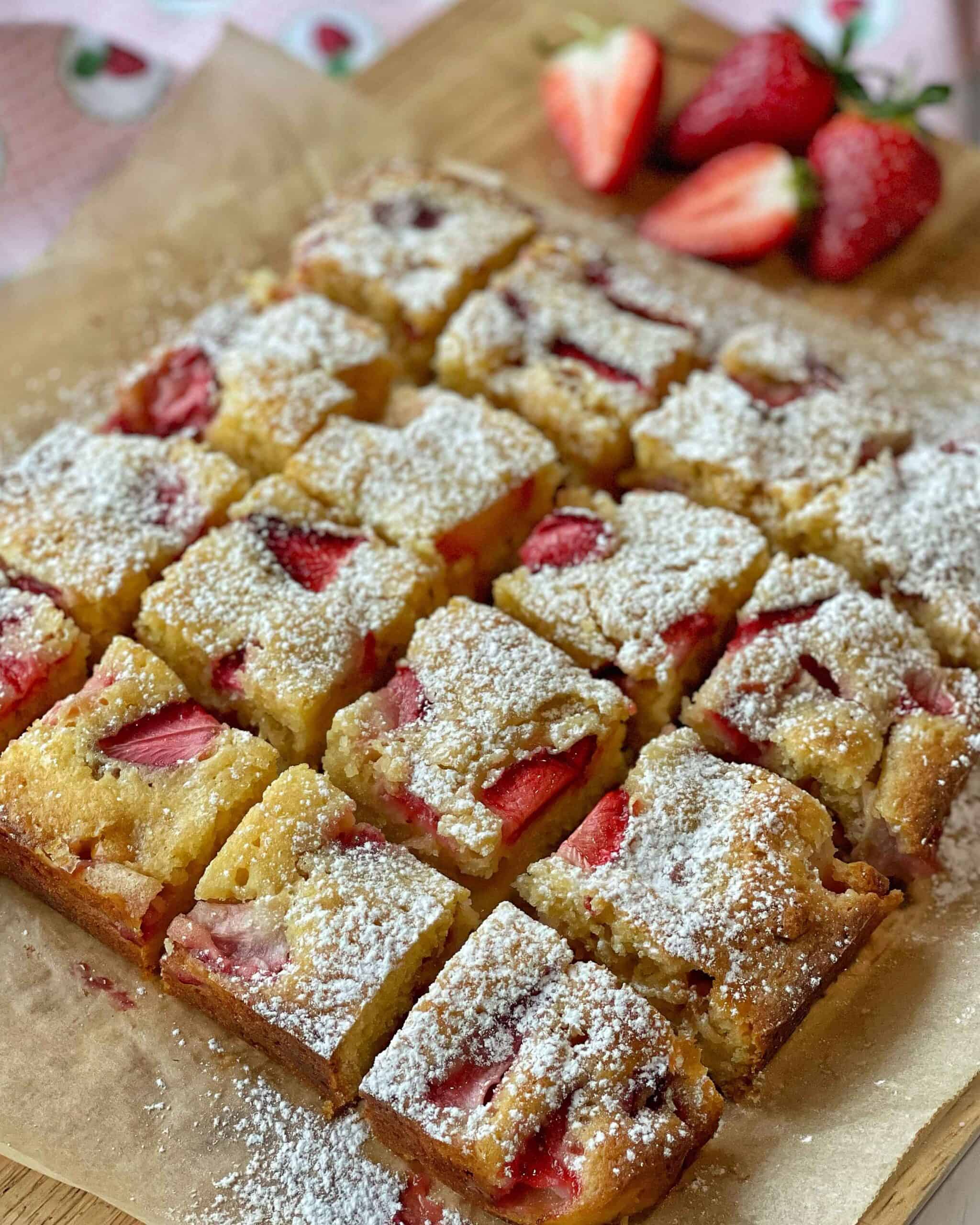 Cooked, sliced White Chocolate and Strawberry Blondie, dusted with icing sugar on baking paper on a chopping board.
