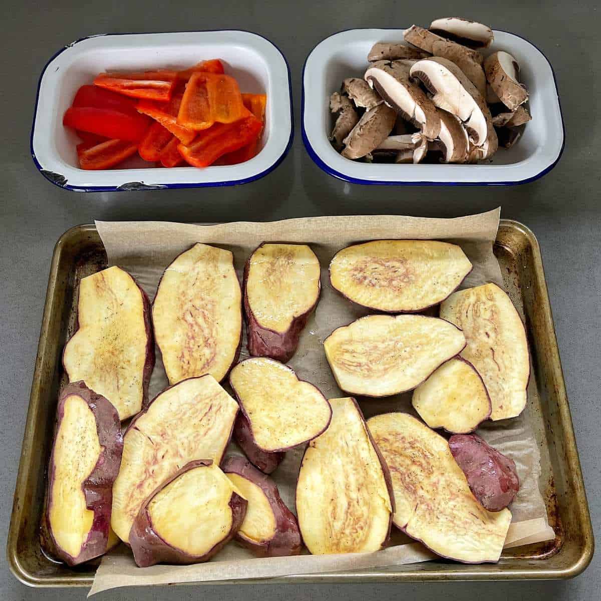 Sliced kumara, large mushrooms and red capsicum in separate dishes on a grey bench top