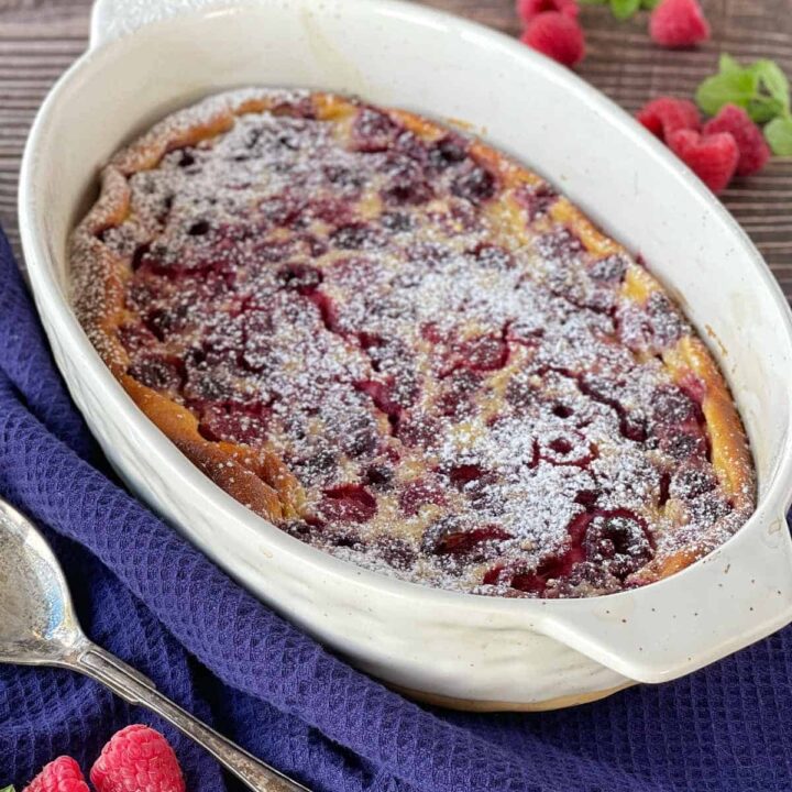 Cooked raspberry calfoutis in a large white oval oven dish sitting on a wooden table with a dark blue napkin
