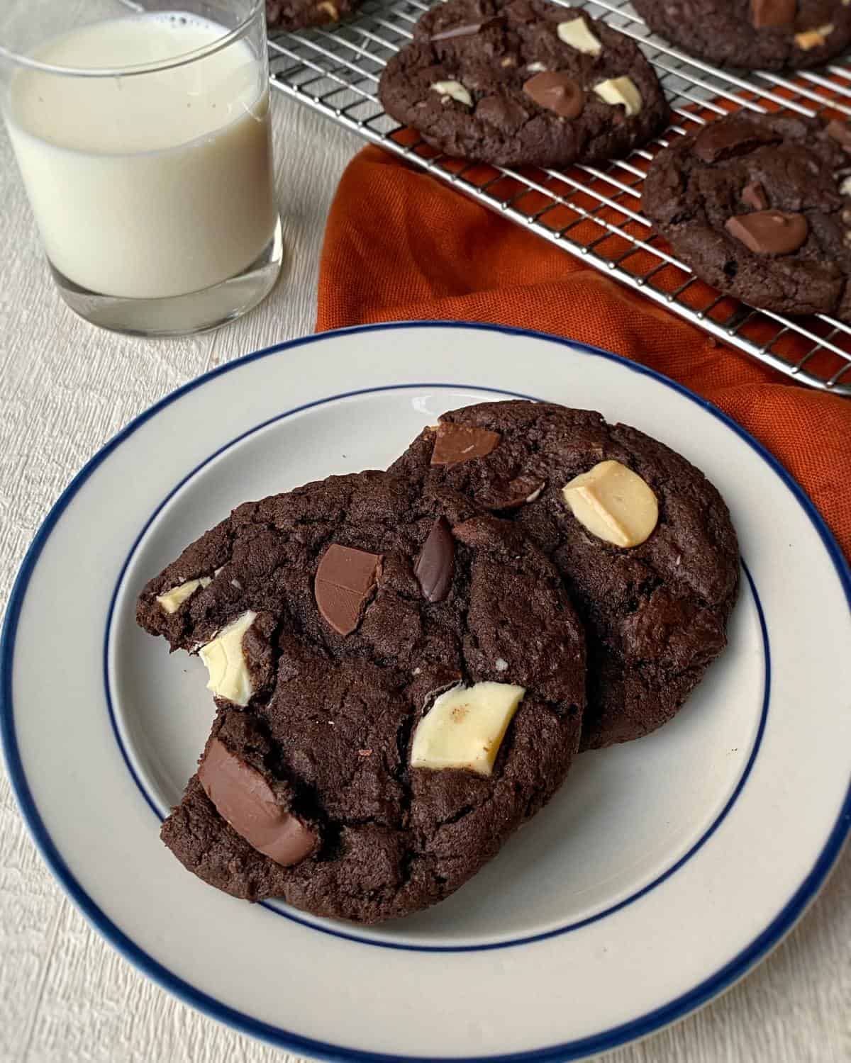 Two triple chocolate chip cookies served on a white plate with a small glass of milk in the background