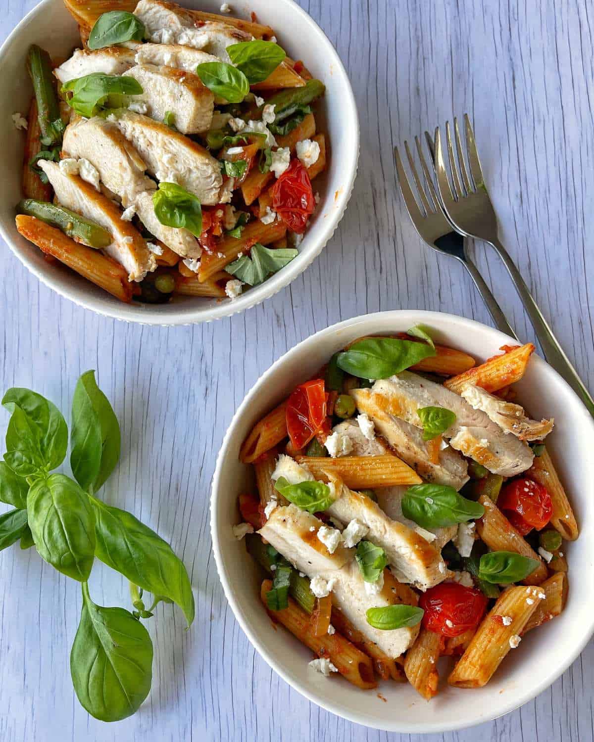 Chicken tomato and asparagus pasta served in a white bowl with scatted basil leaves on top.